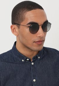 Ochelari de soare rotunzi, negri, cu lentile întunecate, ramă subțire din metal și un logo pe o lentilă. Purtați de o persoană într-o cămașă din denim închis.