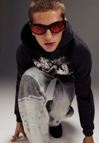 Čierna mikina s grafickým dizajnom, šedé denimové džínsy a prehnane veľké slnečné okuliare s červeným odtieňom. Model pózuje v skrčenej pozícii na šedom pozadí.