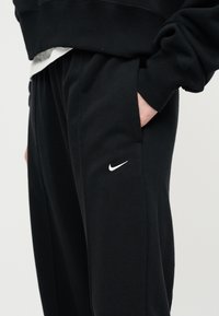 Άτομο που φοράει μαύρο παντελόνι φόρμας Nike με λευκό λογότυπο Swoosh στο μηρό, χέρι στην τσέπη και μαύρο φούτερ εμφανές μερικώς.