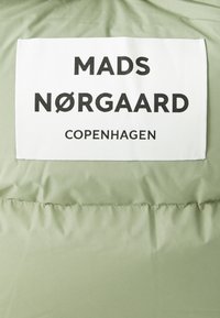 Un tissu de couleur vert clair avec une texture lisse présente une étiquette blanche affichant "MADS NØRGAARD COPENHAGEN" en lettres noires en gras.