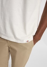 Camiseta blanca de algodón con mangas cortas, un corte holgado y una etiqueta en el dobladillo; combinada con pantalones beige de tejido texturizado.
