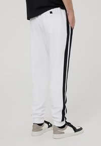 Pantaloni sportivi bianchi con strisce laterali nere, una singola tasca posteriore e polsini a coste, abbinati a sneakers nere e grigie.