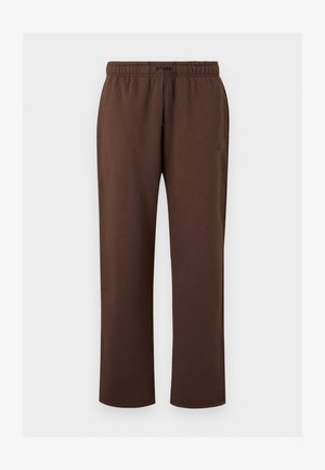 Άτομο που φοράει φαρδιά καφέ φόρμα με ρυθμιζόμενο κορδόνι στη μέση και ανοιχτό γκρι αθλητικά παπούτσια σε απλό φόντο.