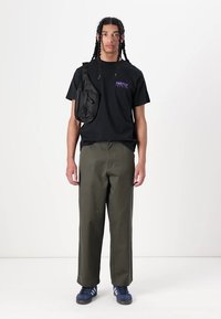 Zwart T-shirt met paarse tekst, olijfgroene wijde broek en marineblauwe sneakers. Een zwarte schoudertas wordt aan de linkerschouder gedragen.