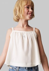 Weißes, strukturiertes Tanktop mit dünnen Trägern, das gefältetes Material und ein kleines "CK"-Logo auf der Vorderseite zeigt und mit Jeansshorts kombiniert wird.
