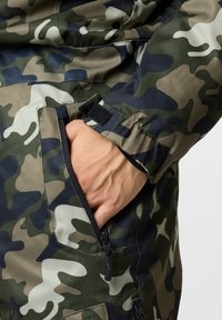 Kamoflaasijakku, jossa on sekoitus vihreitä ja beigejä kuvioita, sekä kankaan tekstuuri näkyvissä. Sisältää vetoketjullisen taskun ja tarranauhakiristykset hihansuissa.