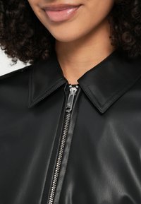 Melna ādas jaka ar asu apkakli, rāvējslēdzēja aizdari un spīdīgu sudraba apdari; gluda tekstūra ar minimālām šuvēm.