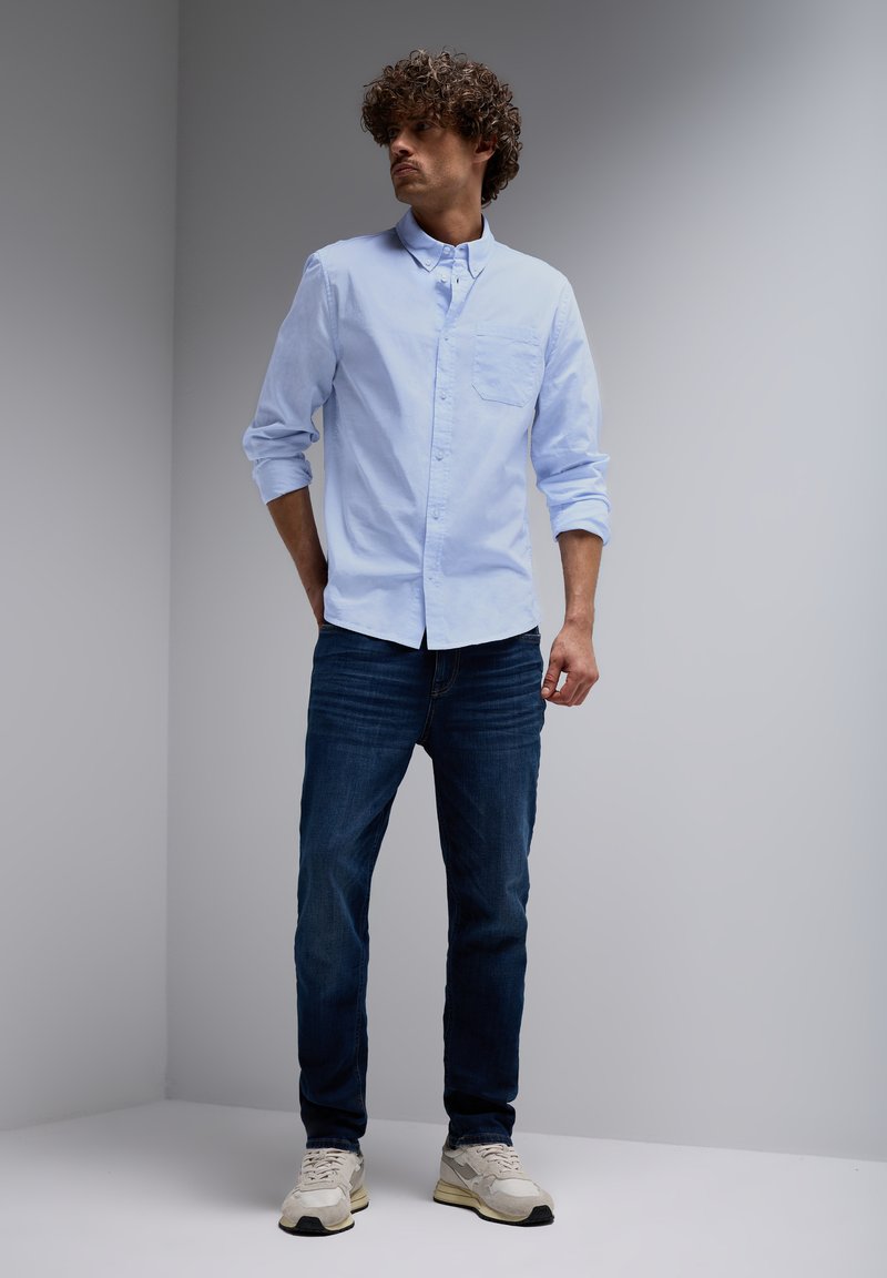 Light blue button-up shirt with a chest pocket, paired with dark blue jeans and light-colored sneakers. Smooth fabric, regular-fit design.