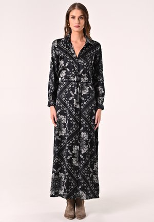 Robe longue à manches longues noire avec un motif floral blanc, taille ceinturée, col et silhouette fluide. Portée avec des bottines brunes.