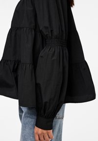 Blusa nera a maniche lunghe con design a strati, dettagli arricciati in vita e maniche a palloncino. Realizzata in tessuto leggero.