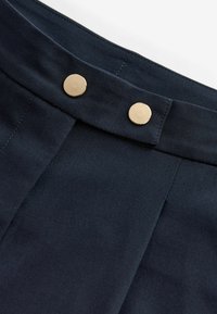 Tessuto blu navy dalla texture liscia presenta due bottoni a pressione dorati in vita, evidenziando un design con pieghe frontali.