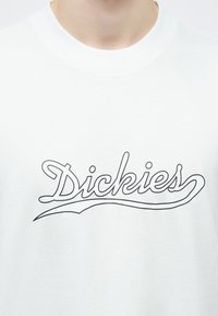 Λευκό μπλουζάκι με στρογγυλή λαιμόκοψη που έχει την λέξη «Dickies» σε μαύρο με περίγραμμα γραφικό κείμενο στο στέρνο.
