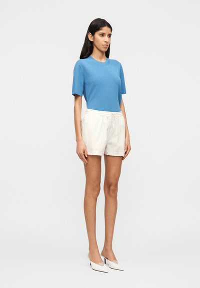 Femme debout portant un haut bleu à manches courtes, un short blanc à cordon avec un motif floral, et des chaussures blanches à talons pointus.