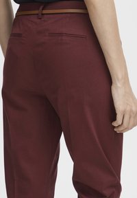 Pantalons bordeaux à la texture lisse, avec des poches arrière et une ceinture marron. Design simple et ajusté.