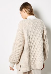 Beige gestrickter Pullover mit strukturiertem Rautenmuster, gerippten Bündchen und Saum, ausgestattet mit einem lockeren Schnitt und seitlichen Schlitzen für Bewegungsfreiheit.