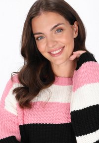 Pulover tricotat cu dungi în roz, alb și negru. Textură ribată cu mâneci largi și guler rotund. Modelul zâmbind și atingându-și bărbia.