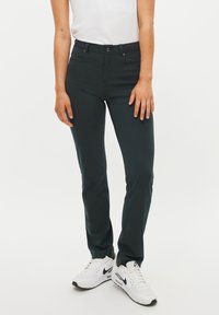 Pantaloni aderenti verde scuro con una texture liscia, cinque tasche e chiusura a bottone, abbinati a sneaker bianche con un logo nero.