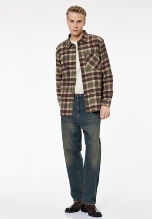 Jeune homme portant une chemise à carreaux verte et marron boutonnée sur un t-shirt blanc, jean bleu délavé et bottes en cuir marron foncé, debout devant un fond blanc.