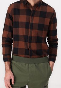 Hnědá a černá kostkovaná košile s dlouhými rukávy a zapínáním na knoflíky; nošena se zelenými kalhotami s elastickým pasem. Hladká textilie.