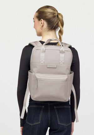 Frau mit blondem Zopf, die einen hellgrauen Rucksack mit vorderer Reißverschlusstasche und Seitentaschen über einem schwarzen Langarmshirt und Jeans trägt.