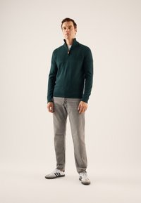 Dunkelgrüner Strickpullover mit hohem Kragen, ausgestattet mit einem halben Reißverschluss und strukturiertem Muster. Kombiniert mit hellgrauen Jeans und schwarz-weißen Sneakers.