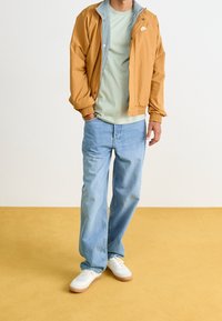 Tansfärgad bomberjacka med grå innertyg, ljusgrön t-shirt, blå raka jeans och vita sneakers med gummisula.