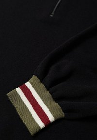 Pull noir à col zippé et tricot texturé. Les poignets ont une base verte avec des rayures rouges et blanches.