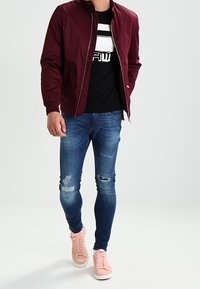 Burgundská bomber bunda přes černé tričko s bílou grafikou, kombinovaná s modrými skinny džíny s dírami na kolenou a světle růžovými teniskami.