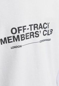Bílá bavlněná tričko s velkým černým textem: "OFF-TRACK MEMBERS' CLUB," se sídlem "LONDÝN" a "KODAŇ" níže.