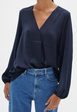 Kvinde iført en mørkeblå, langærmet V-hals bluse, der er stoppet ned i en højtaljet, midtblå jeans med knappet linning.