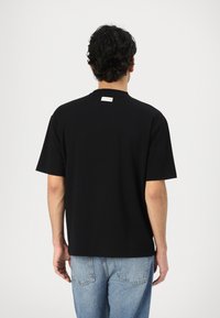 Black short-sleeve T-shirt with a crew neck, made from cotton, features a small brand label at the back neck. Worn with light blue jeans.