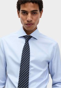 Mężczyzna z kręconymi włosami, ubrany w jasnoniebieską koszulę i ciemnoniebieski pasiasty krawat, zwrócony przodem na jednolitym tle.