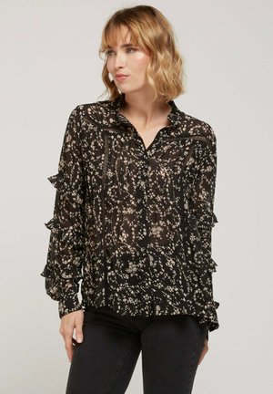 Blouse noire à fleurs avec manches longues, accents volantés, devant boutonné et en tissu transparent. Présente de petits motifs floraux clairs.