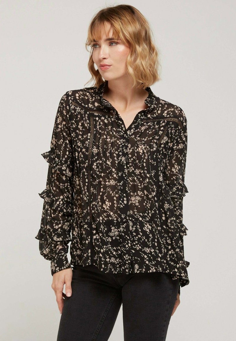 Blouse noire à fleurs avec manches longues, accents volantés, devant boutonné et en tissu transparent. Présente de petits motifs floraux clairs.