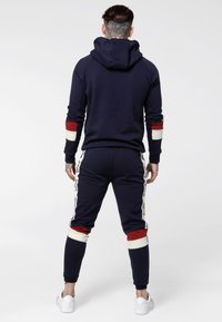 Chándal azul marino con capucha, con acentos de rayas rojas y blancas en las mangas y los pantalones, confeccionado en tejido suave. Zapatillas blancas incluidas.