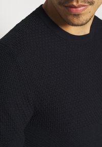 Närbild av en man som bär en mörk, strukturerad stickad tröja med rund halsringning, visar läppar, haka och en del av axeln.