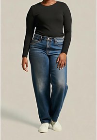 Personne portant une chemise noire à manches longues, un jean bleu avec une braguette à boutons et des chaussures blanches sans lacets, debout sur un fond uni.