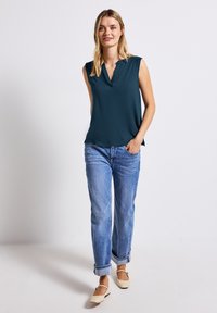 Ciemnoniebieska bezrękawnik z dekoltem w serek o gładkiej fakturze, zestawiona z jasnoniebieskimi dżinsami i beżowymi płaskimi butami, stojąca na jasnym tle.