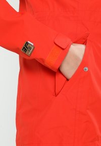 Main dans la poche d'une veste orange vif avec une bride Velcro au poignet et un petit patch marron cousu près du poignet.