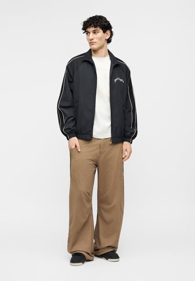 Nooruk lokkis juustega, kandes musta lukuga jopet valge ääristusega, valget t-särki, laiu pruune pükse ja musti tossusid, seismas.