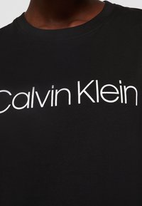Černé bavlněné tričko s velkým bílým logem "Calvin Klein" na hrudi, s kulatým výstřihem a krátkými rukávy. Hladká textura.