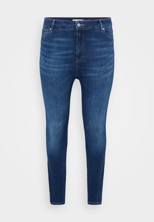 Jean skinny bleu foncé avec poches avant, passants de ceinture et fermeture à bouton sur un fond uni clair.