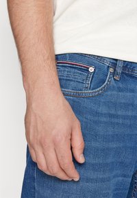 Blå denimjeans med en figurnära passform, med röd och vit sömdetalj längs fickan, knappmidja och en lätt texturvariation.