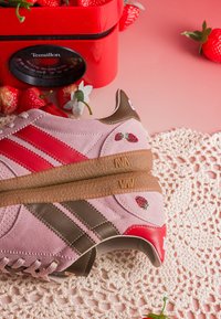 Paire de baskets en daim rose avec des rayures rouges et marron, petite broderie de fraise, placées à côté d'une balance de cuisine rouge et de fraises fraîches.