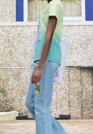 Osoba w koszulce z gradientem od zielonego do niebieskiego i jasnoniebieskich dżinsach z kolorową naszywką, niosąca białą torbę na ramię, spacerująca na zewnątrz przy teksturowanej ścianie.