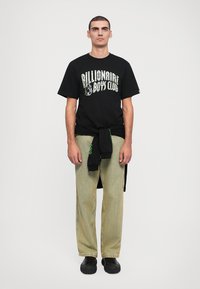Schwarzes T-Shirt mit dem Logo "Billionaire Boys Club", grünen Akzenten, kombiniert mit beigen gestreiften Hosen. Schlichte schwarze Schuhe. Minimalistischer Stil.