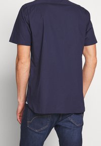 Uomo che indossa una camicia a maniche corte di colore blu navy e jeans blu scuri, visto di spalle su uno sfondo neutro.