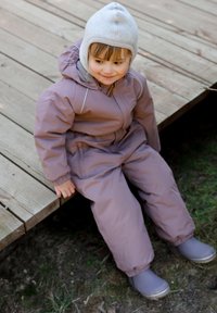 Lapsi vaaleanpunaisessa toppahaalarissa, jossa on harmaat yksityiskohdat ja harmaa hattu, istuu puualustalla ja pukeutuneena harmaisiin saappaisiin.