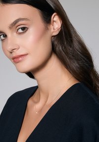 Złota naszyjnik łańcuszkowy z małym, okrągłym wisiorkiem. Noszony z głębokim dekoltem w kształcie V. Gładkie włosy opadające na ramię, neutralne tło.