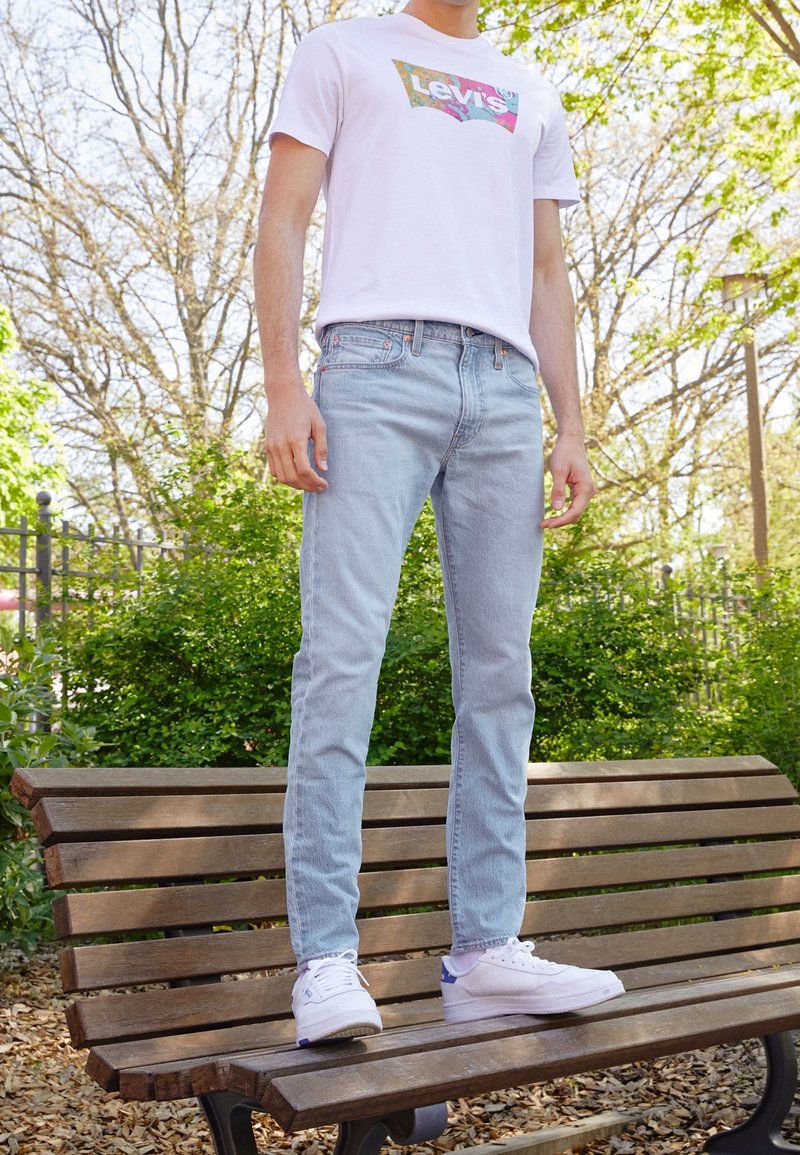 Vit t-shirt med färggloggig Levi's-logotyp, ljusblå denimjeans och vita sneakers med lila detaljer. Utomhus, stående på en träbänk.